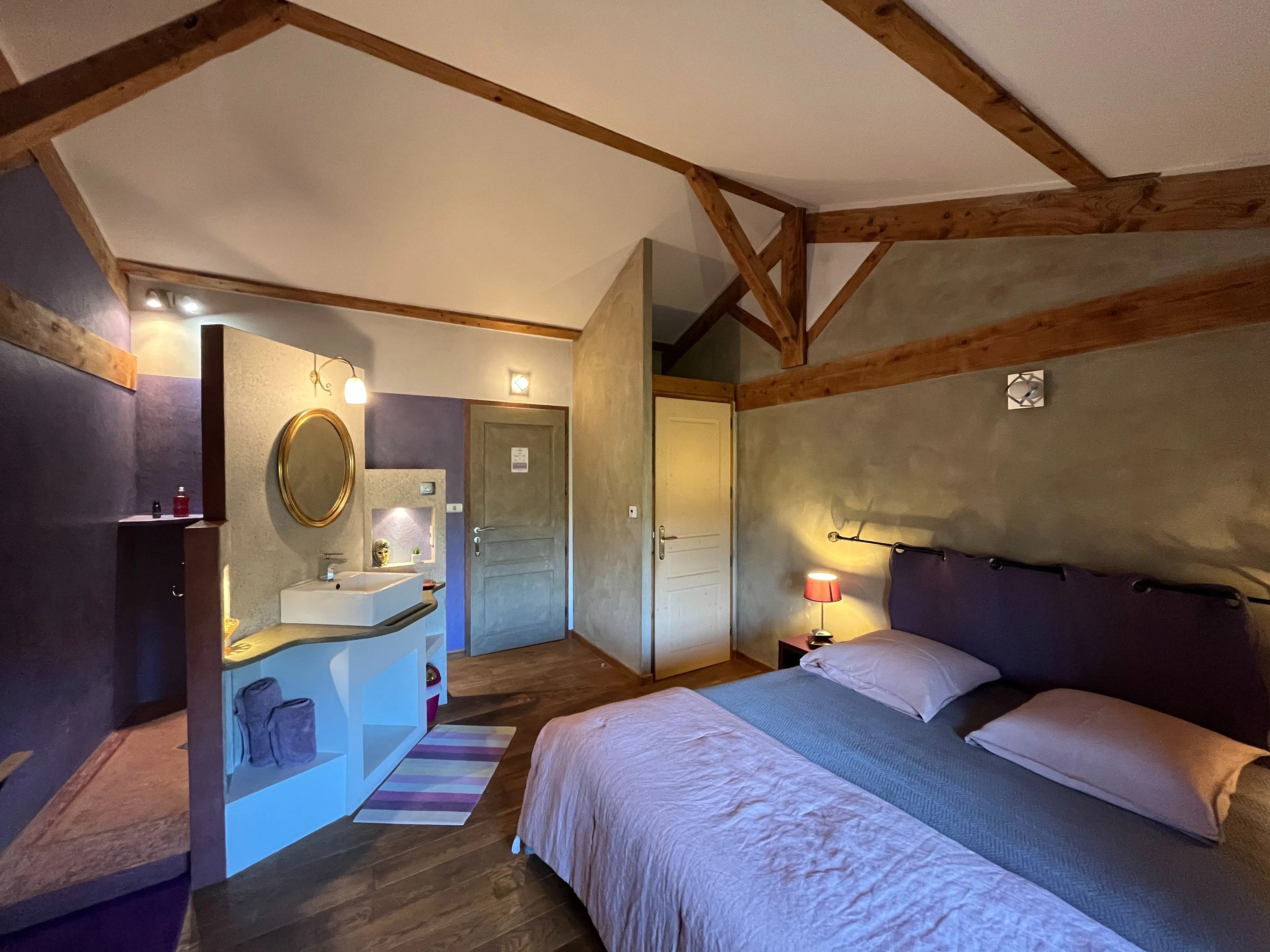 Chambre toscane - Vue d’ensemble de la chambre avec lit double et lampe de chevet allumée, entrée centrale, porte des toilettes et grande douche en tadelakt avec vasque simple